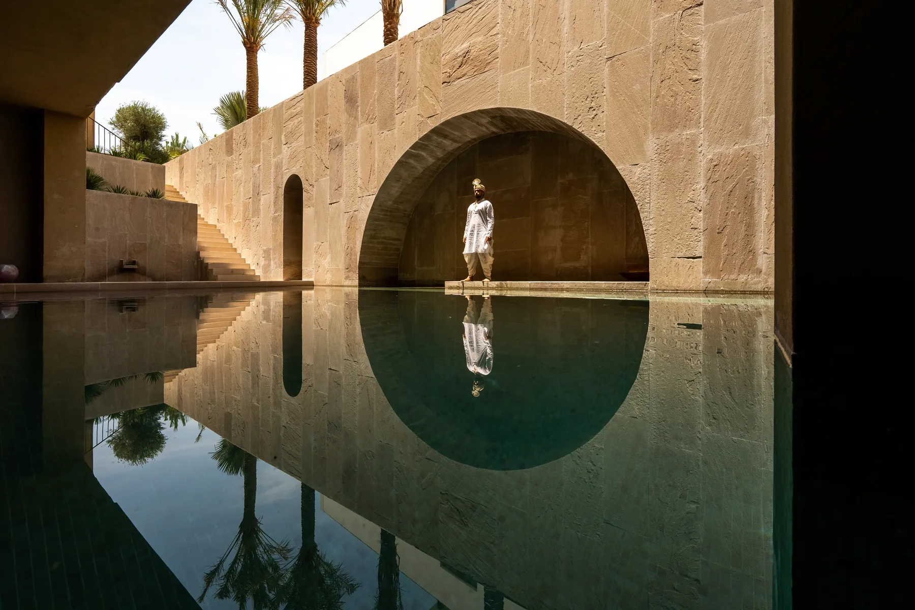 A figure standing in the arched alcove at the end of the courtyard pool, mirrored in still water