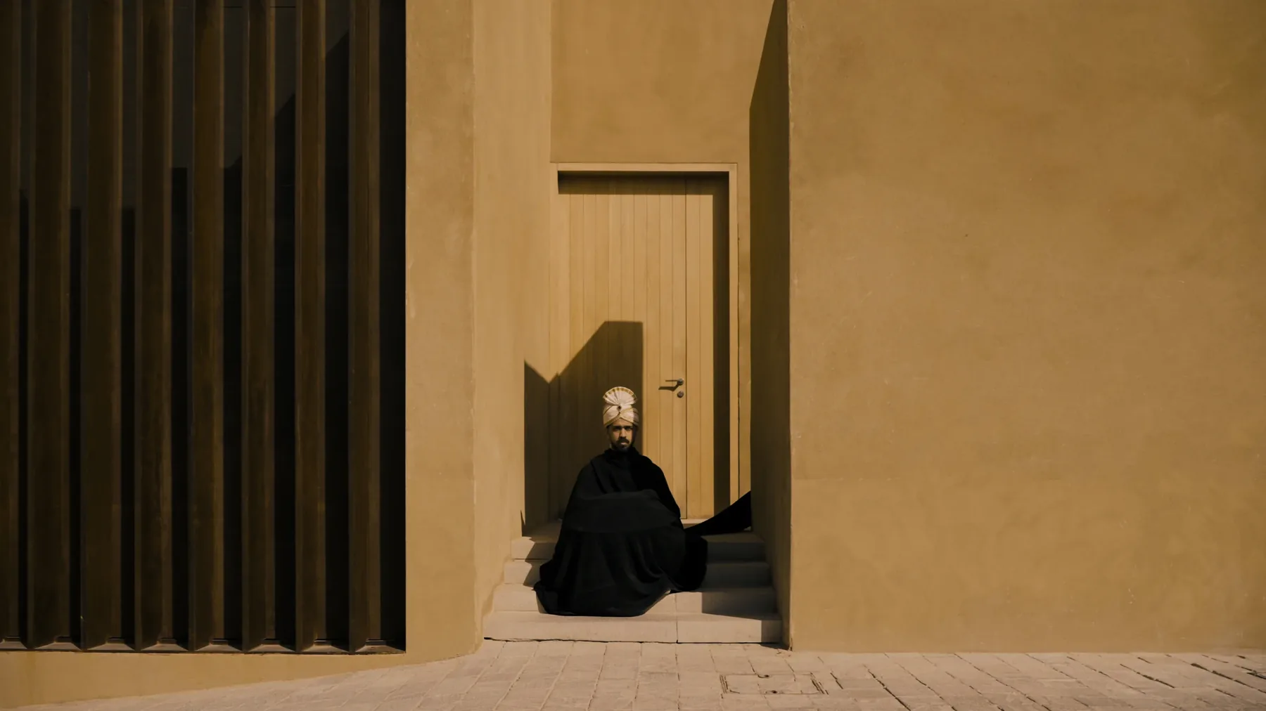 A seated figure in black framed by a doorway between fluted and plain ochre walls